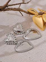 Load image into Gallery viewer, These romantic drop earrings feature an enchanting design with a larger heart gracefully dangling from a smaller one, each crafted from hypoallergenic stainless steel and plated in radiant 18K gold or classic silver. The textured surface and intricate edges add depth and charm, creating an eye-catching look that's both elegant and playful.