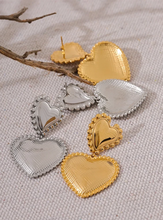 Load image into Gallery viewer, These romantic drop earrings feature an enchanting design with a larger heart gracefully dangling from a smaller one, each crafted from hypoallergenic stainless steel and plated in radiant 18K gold or classic silver. The textured surface and intricate edges add depth and charm, creating an eye-catching look that's both elegant and playful.