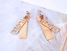 Load image into Gallery viewer, These fashionable stainless steel earrings are made in a rose gold colour or combination of silver and rose gold. They consists of a sanded rectangle and letters LOVE. Both, letters and rectangle are hanging on a circle.Earrings are three times gold plated and represent perfect and elegant touch to your day to day style. They have stud fastening.
