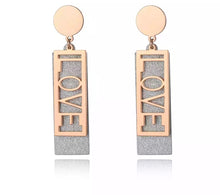 Load image into Gallery viewer, These fashionable stainless steel earrings are made in a rose gold colour or combination of silver and rose gold. They consists of a sanded rectangle and letters LOVE. Both, letters and rectangle are hanging on a circle.Earrings are three times gold plated and represent perfect and elegant touch to your day to day style. They have stud fastening.
