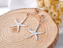 Load image into Gallery viewer, Trendy stainless steel earrings are made in a rose gold colour. They are adorned with starfish that comes in white or black colour. Starfish is made of cubic zirconia and it hangs on a little ball. Earrings are rose gold plated and made great summer accessory. They have stud fastening.
